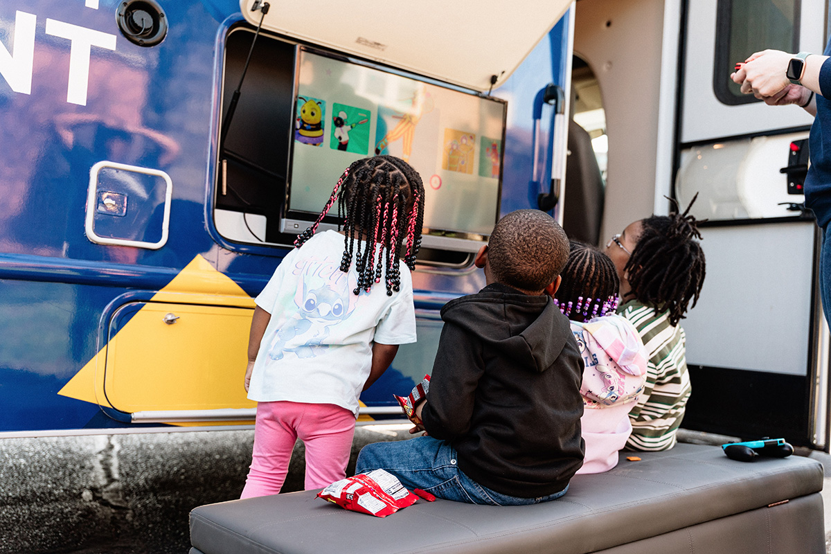 Four children watching a screen mounted on the side of a vehicle, with one child standing and three sitting on a bench.