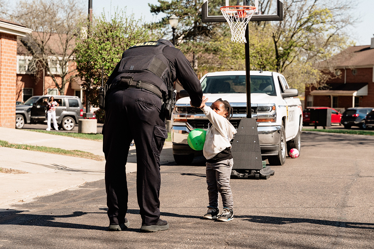 A police officer giving a high-five to a young child holding a green ball near a basketball hoop in a residential area.