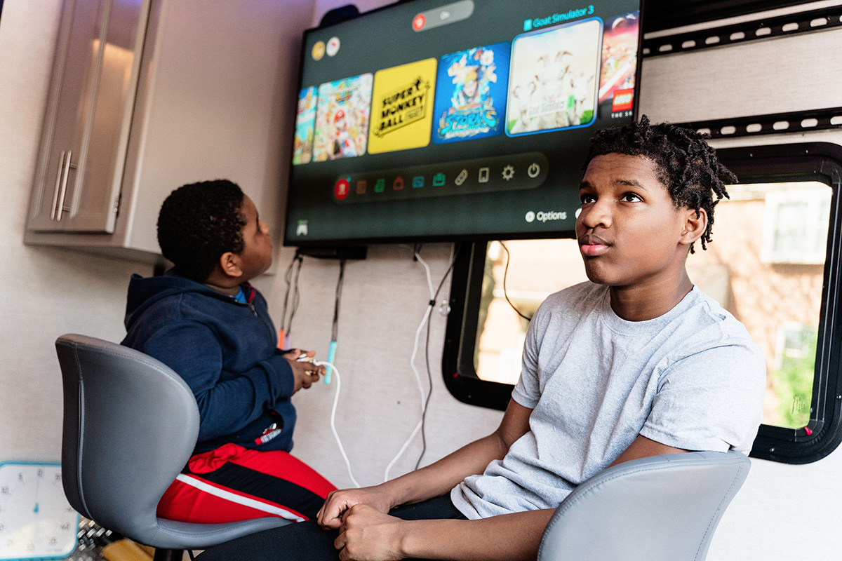 Two boys sitting on chairs playing video games on a wall-mounted TV inside a room.