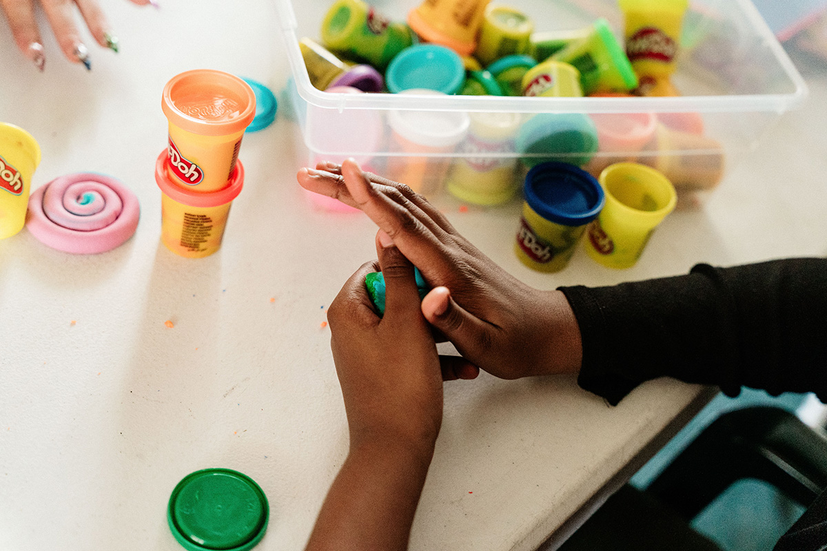 Child's hands molding green Play-Doh on a table with various Play-Doh containers and a pink spiral shape nearby.