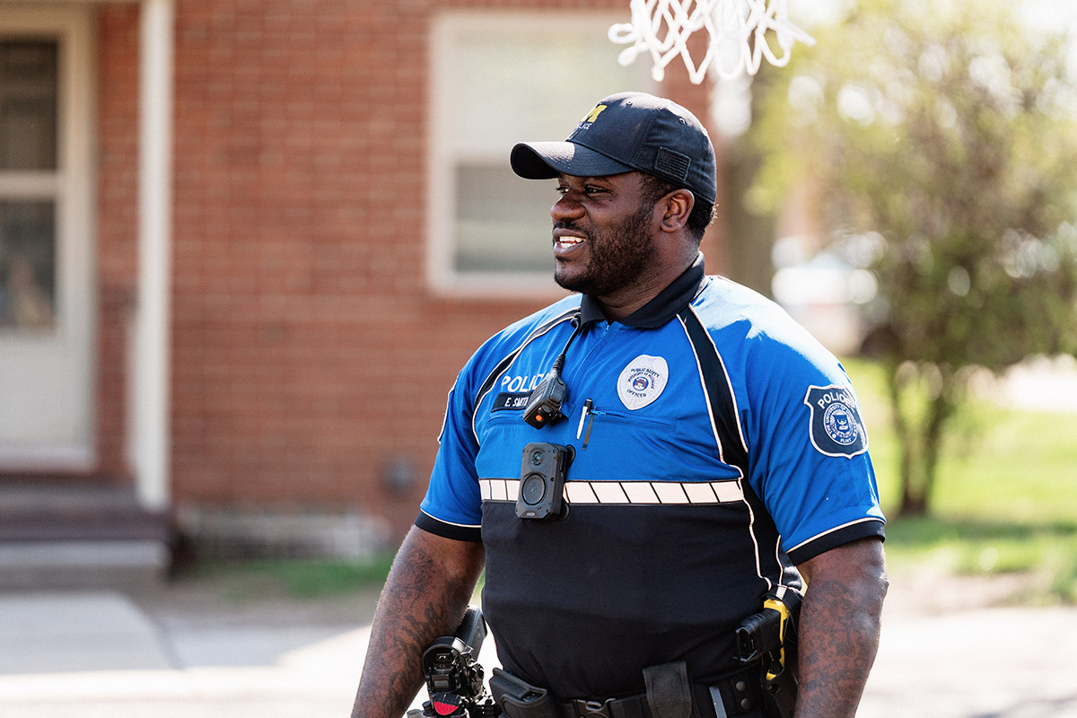 A police officer in a blue and black uniform with a body camera and radio standing outdoors near a basketball hoop.