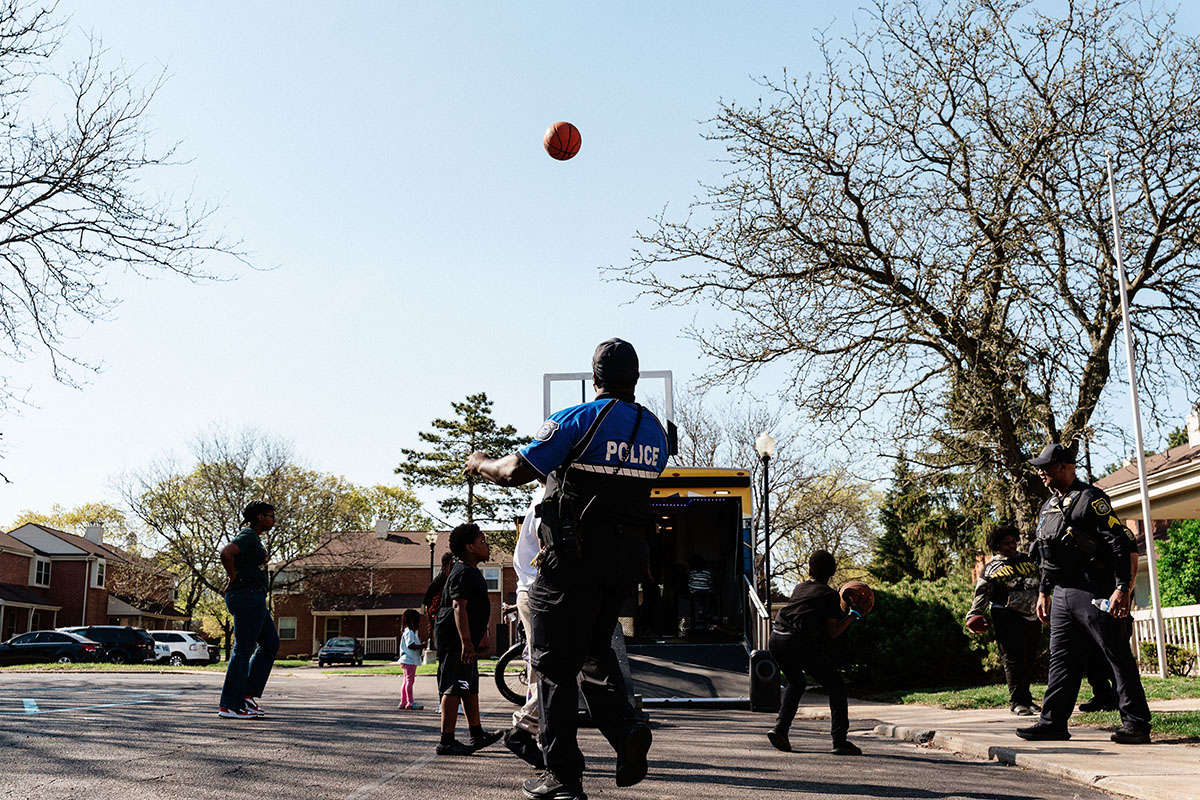 Police officers and children playing basketball outdoors in a neighborhood setting.