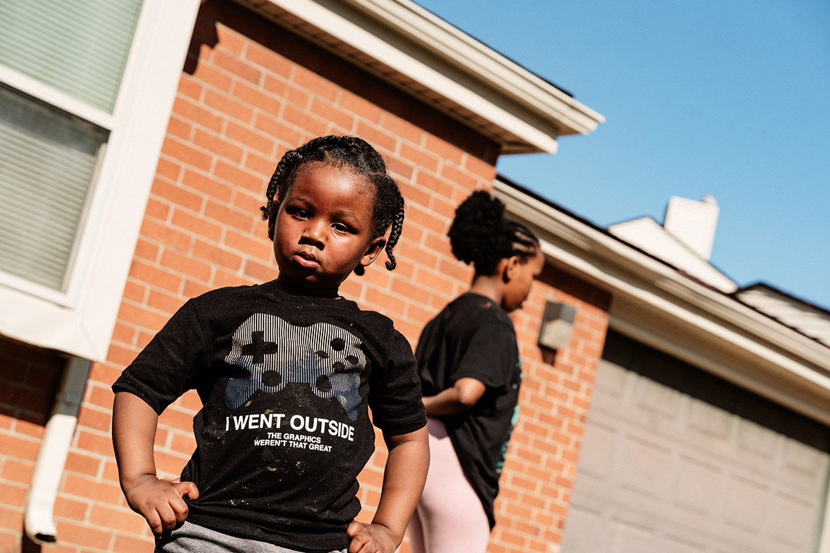Two children standing outside a brick house, one wearing a black shirt with a game controller graphic and the text "I WENT OUTSIDE THE GRAPHICS WEREN'T THAT GREAT"