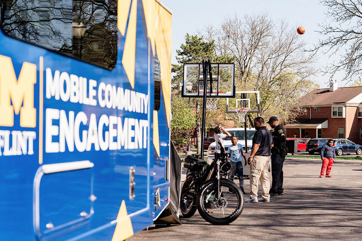 A Mobile Community Engagement vehicle is parked near a basketball hoop where two children and two adults, including a police officer, are gathered, with one child shooting a basketball.
