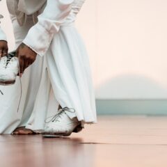 Person in white pants and shirt tying the laces of one white shoe while the other foot is bare.