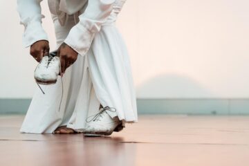 Person in white pants and shirt tying the laces of one white shoe while the other foot is bare.