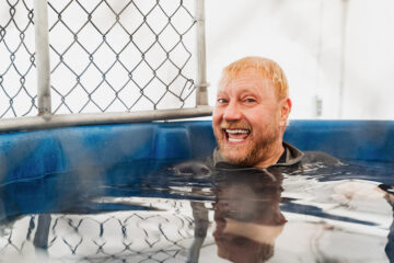 Person with blond hair relaxing in a blue hot tub with a chain-link fence in the background.
