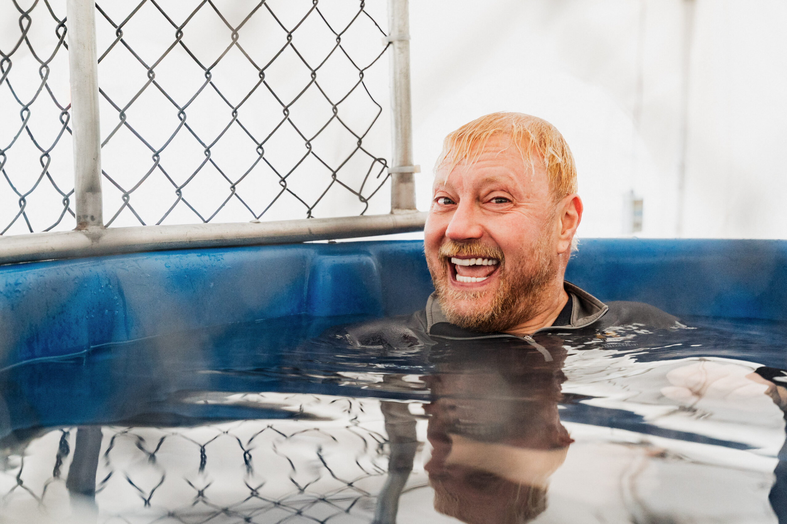 Person with blond hair relaxing in a blue hot tub with a chain-link fence in the background.