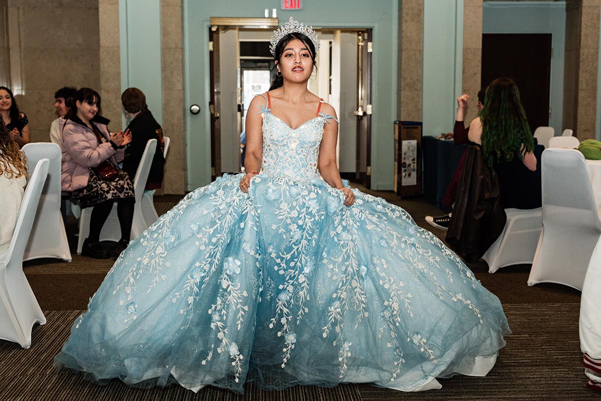 FLINT_LUNA Quinceañera Night 2026_0008 Young woman in a blue floral ball gown and tiara standing in a room with seated guests.