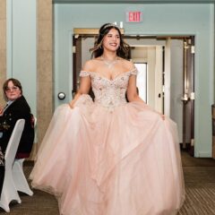 Young woman in an embellished off-shoulder pink ball gown holding the skirt, walking indoors with people seated and clapping on the sides.