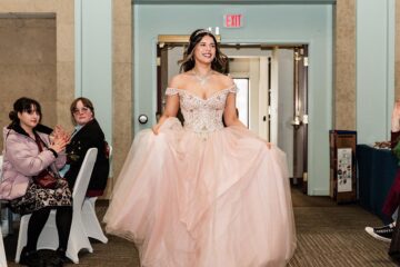 Young woman in an embellished off-shoulder pink ball gown holding the skirt, walking indoors with people seated and clapping on the sides.