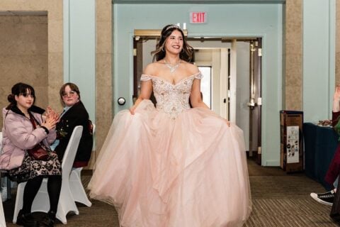Young woman in an embellished off-shoulder pink ball gown holding the skirt, walking indoors with people seated and clapping on the sides.