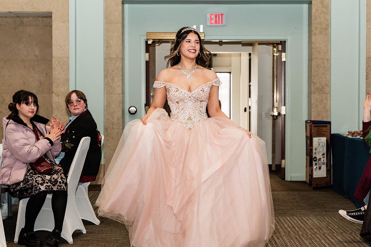 Young woman in an embellished off-shoulder pink ball gown holding the skirt, walking indoors with people seated and clapping on the sides.