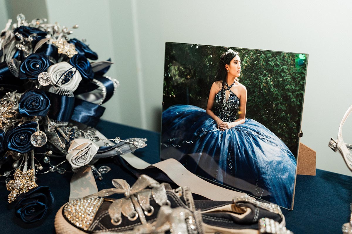 FLINT_LUNA Quinceañera Night 2026_0023 Photo of a young woman in a blue ball gown displayed on a table with a bouquet of blue and white flowers and a pair of black sneakers decorated with rhinestones.