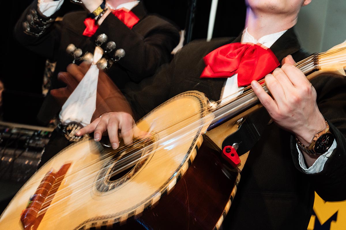 FLINT_LUNA Quinceañera Night 2026_0035 Close-up of a musician wearing a black suit and red bow tie playing a guitar-like instrument with motion blur on the hands.