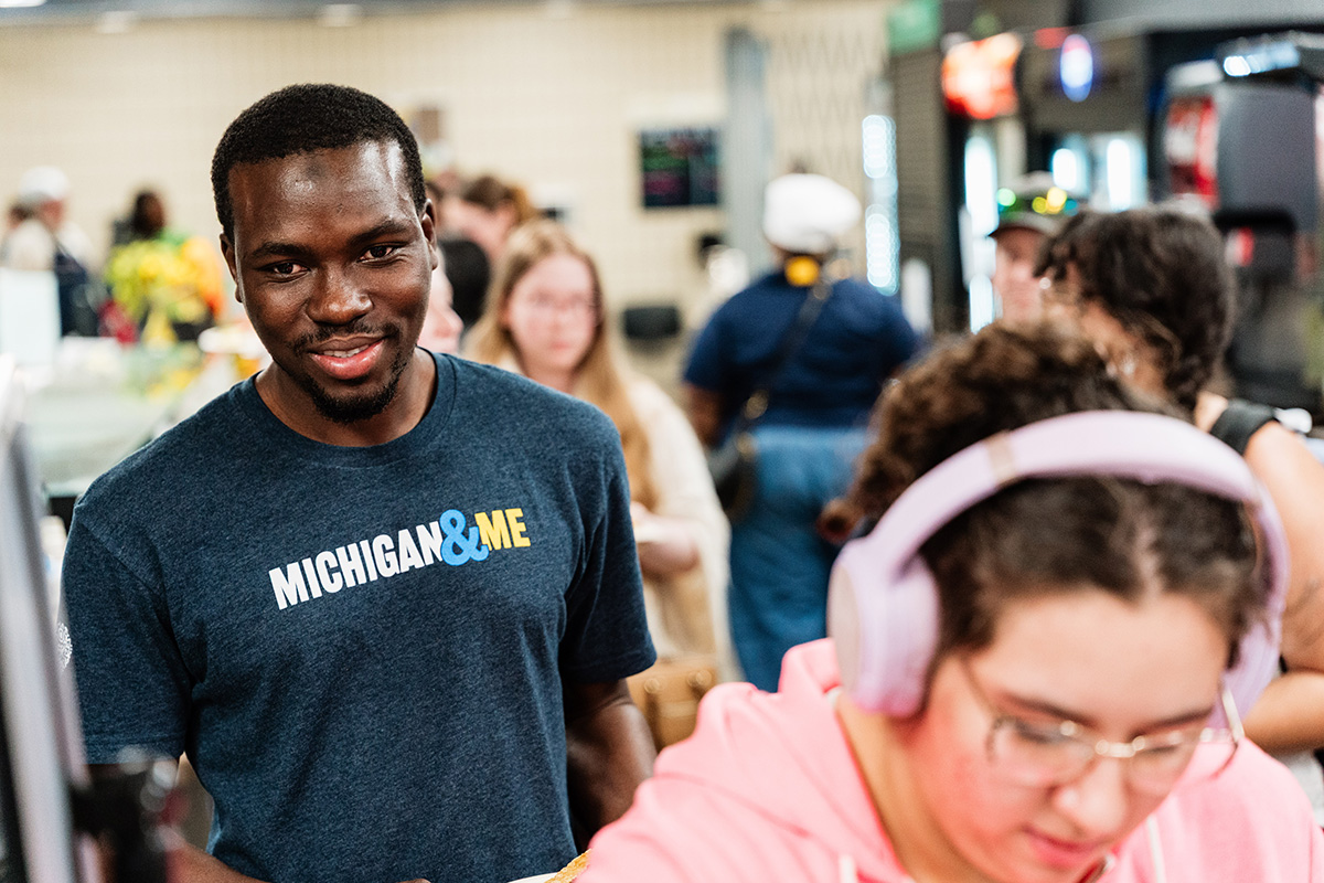 FLINT_Late Night Breakfast 2026_0005 Person wearing a navy blue "MICHIGAN&ME" t-shirt standing in a busy indoor setting.