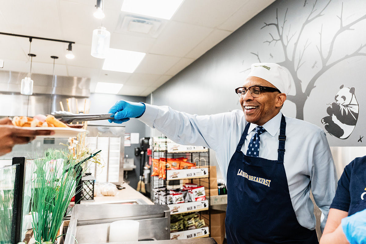 FLINT_Late Night Breakfast 2026_0011 Man wearing a "Lansing Breakfast" apron and gloves serving food with tongs in a kitchen with a panda mural on the wall.