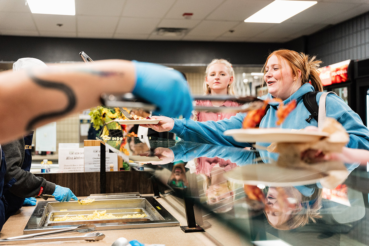 FLINT_Late Night Breakfast 2026_0014 People being served food in a cafeteria line with a person wearing blue gloves using tongs to serve food.