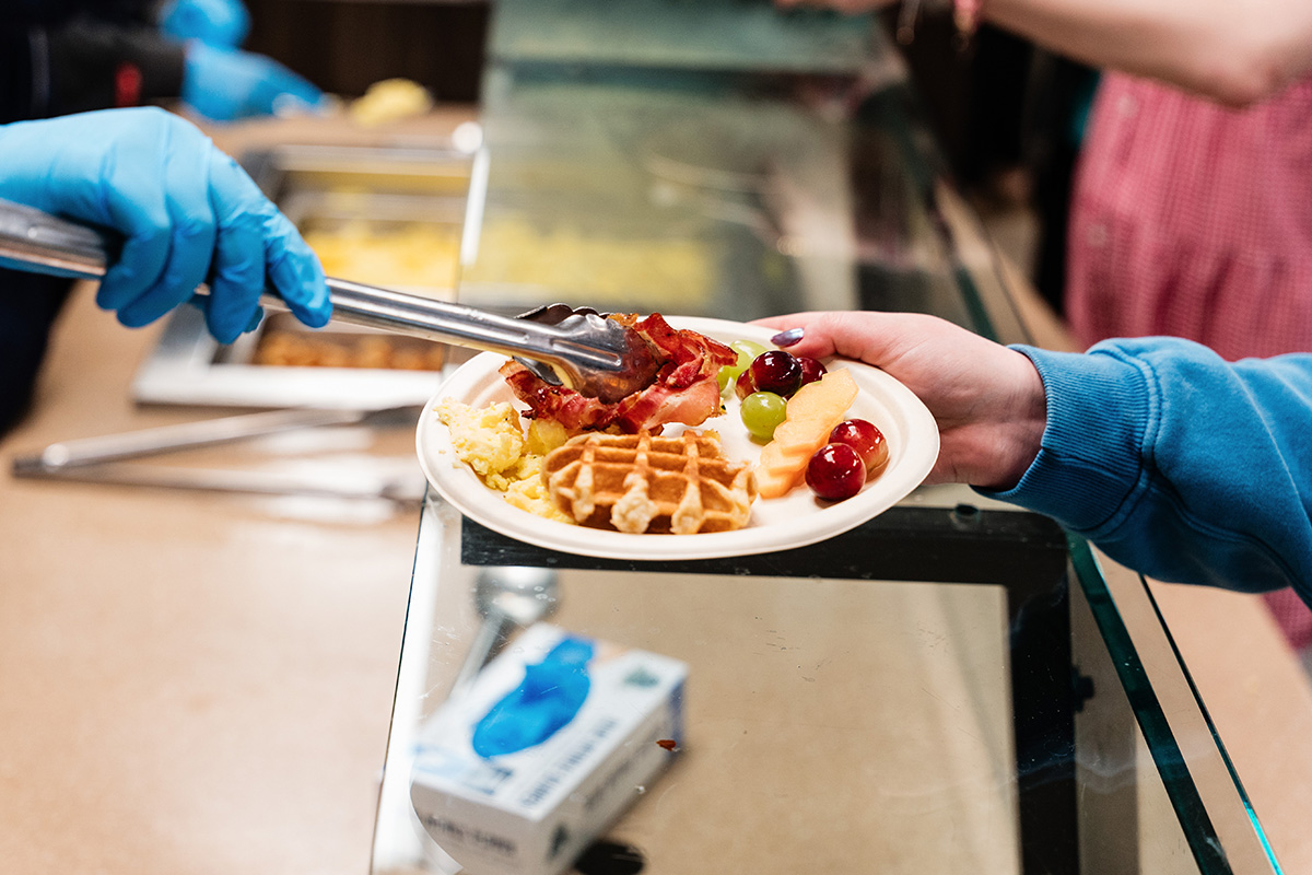 FLINT_Late Night Breakfast 2026_0015 A person wearing a blue glove uses tongs to place bacon on a plate with scrambled eggs, a waffle, and fruit.