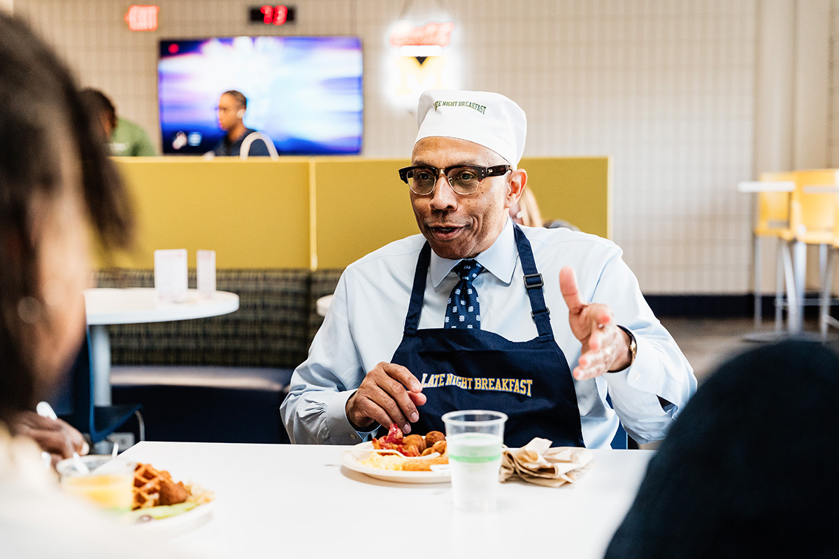 FLINT_Late Night Breakfast 2026_0029 Person wearing a "Late Night Breakfast" apron and hat, sitting at a table with breakfast food, gesturing with one hand.