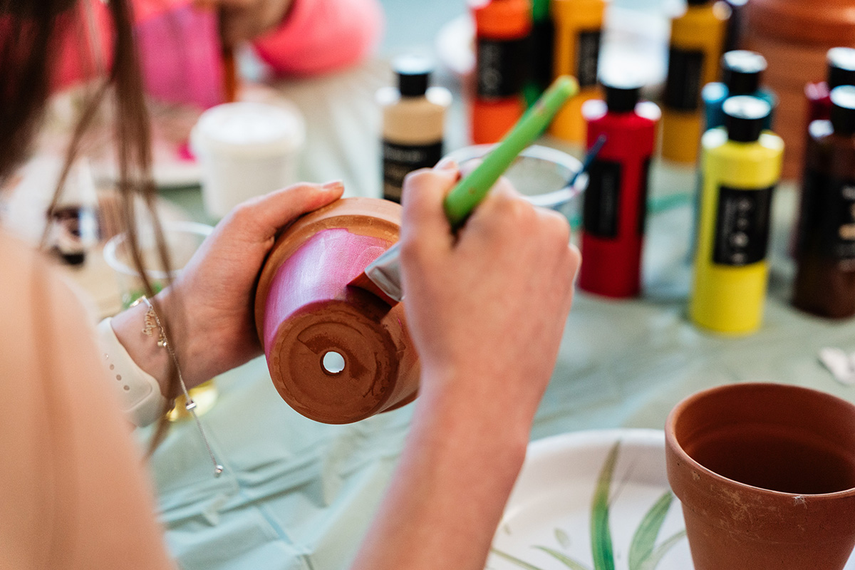FLINT_Late Night Breakfast 2026_0035 Person painting a terracotta pot with pink paint using a brush, with bottles of paint in the background.
