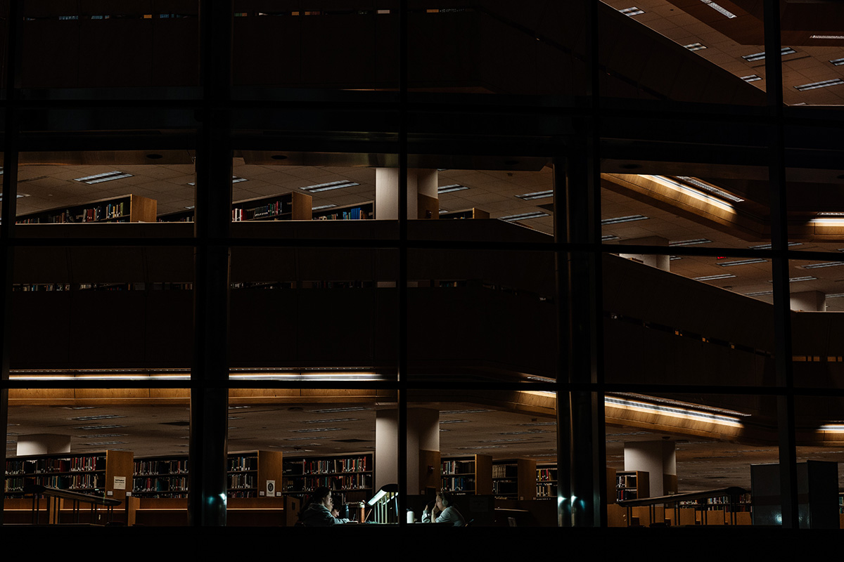 FLINT_Late Night in the Library 2026_0001 View through large windows into a dimly lit library interior with bookshelves and two people studying at a table with a desk lamp.