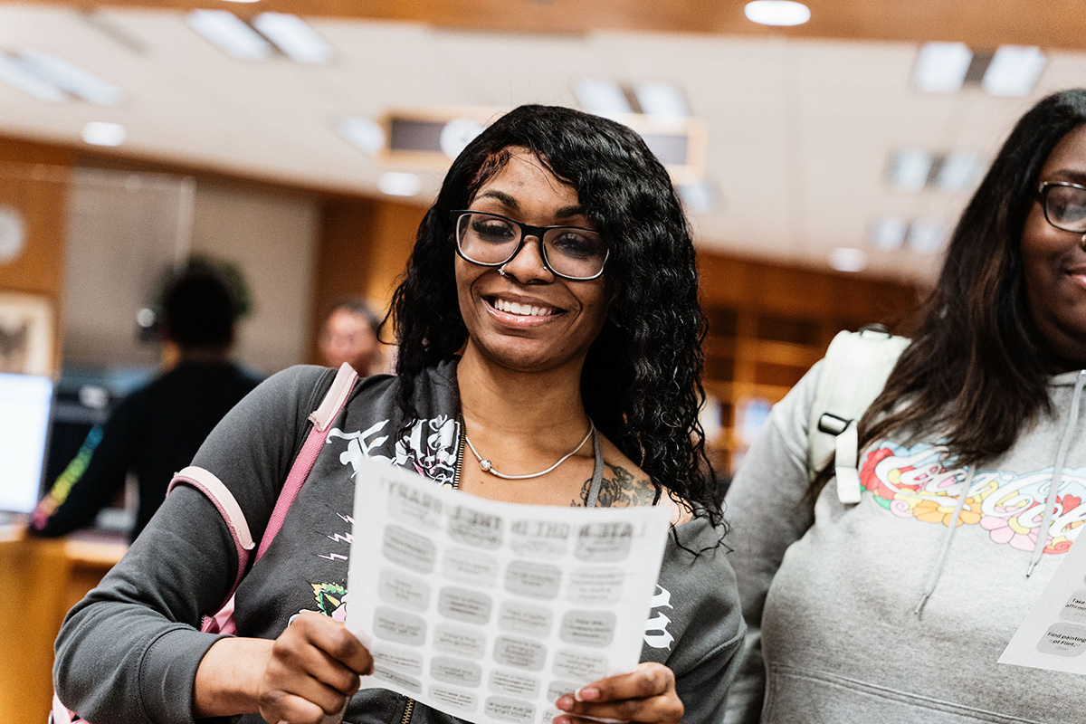 FLINT_Late Night in the Library 2026_0002 Two women standing indoors, one holding a paper with text, both wearing casual clothes and backpacks.