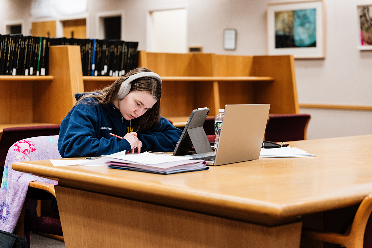 FLINT_Late Night in the Library 2026_0004 Person wearing headphones writing in a notebook at a library table with a laptop, tablet, and water bottle.