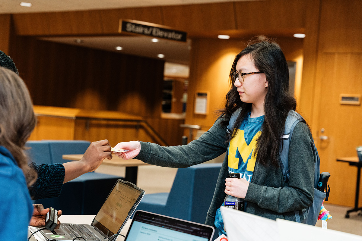 FLINT_Late Night in the Library 2026_0008 A person wearing a University of Michigan shirt and backpack hands a card to another person across a table with laptops in a wood-paneled room.