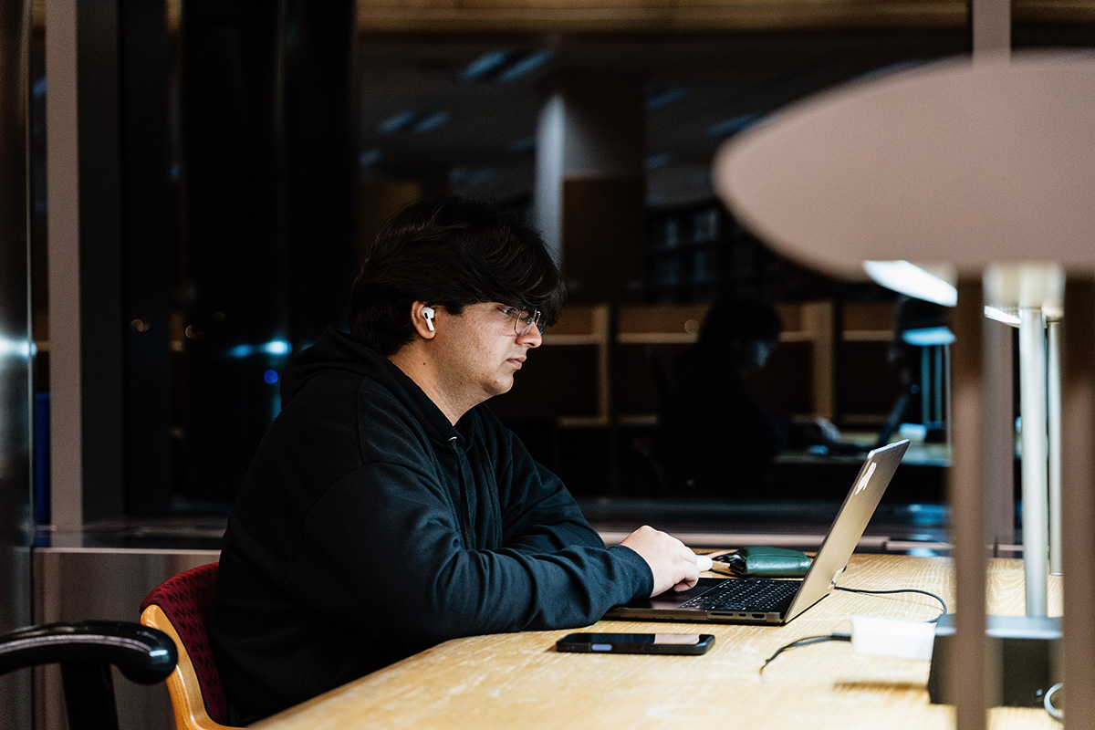 FLINT_Late Night in the Library 2026_0013 Person wearing a black hoodie and white wireless earbuds working on a laptop at a wooden table in a dimly lit room.