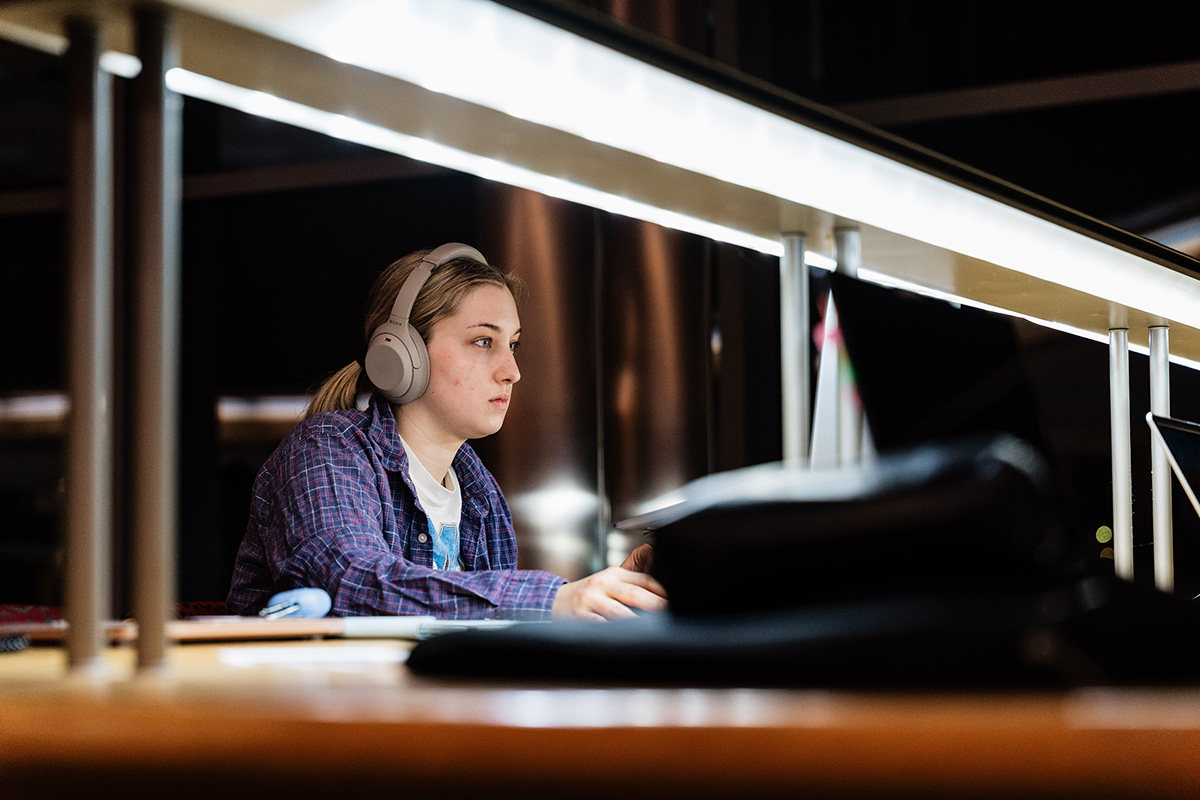 FLINT_Late Night in the Library 2026_0015 Person wearing headphones and a plaid shirt working on a laptop at a desk with overhead lighting.