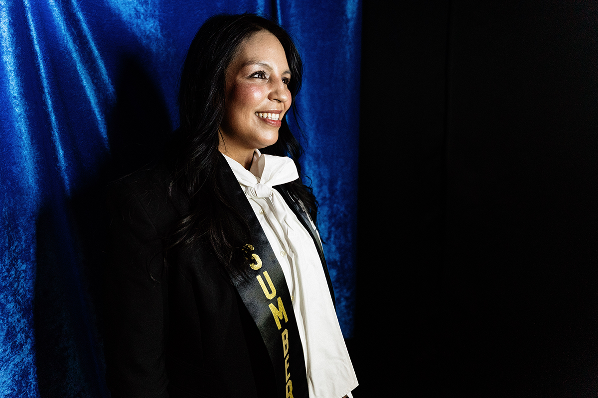 FLINT_Latin Graduation Celebration 2026_0010 Person wearing a black jacket, white blouse with a bow, and a black sash with gold letters reading "NUMBER" against a blue and black background.