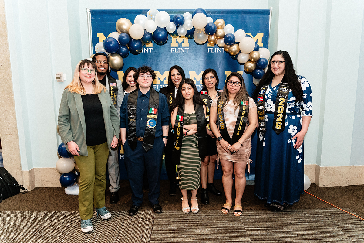 FLINT_Latin Graduation Celebration 2026_0025 Group of eight people standing in front of a University of Michigan-Flint backdrop with a blue, white, and gold balloon arch, some wearing graduation stoles.