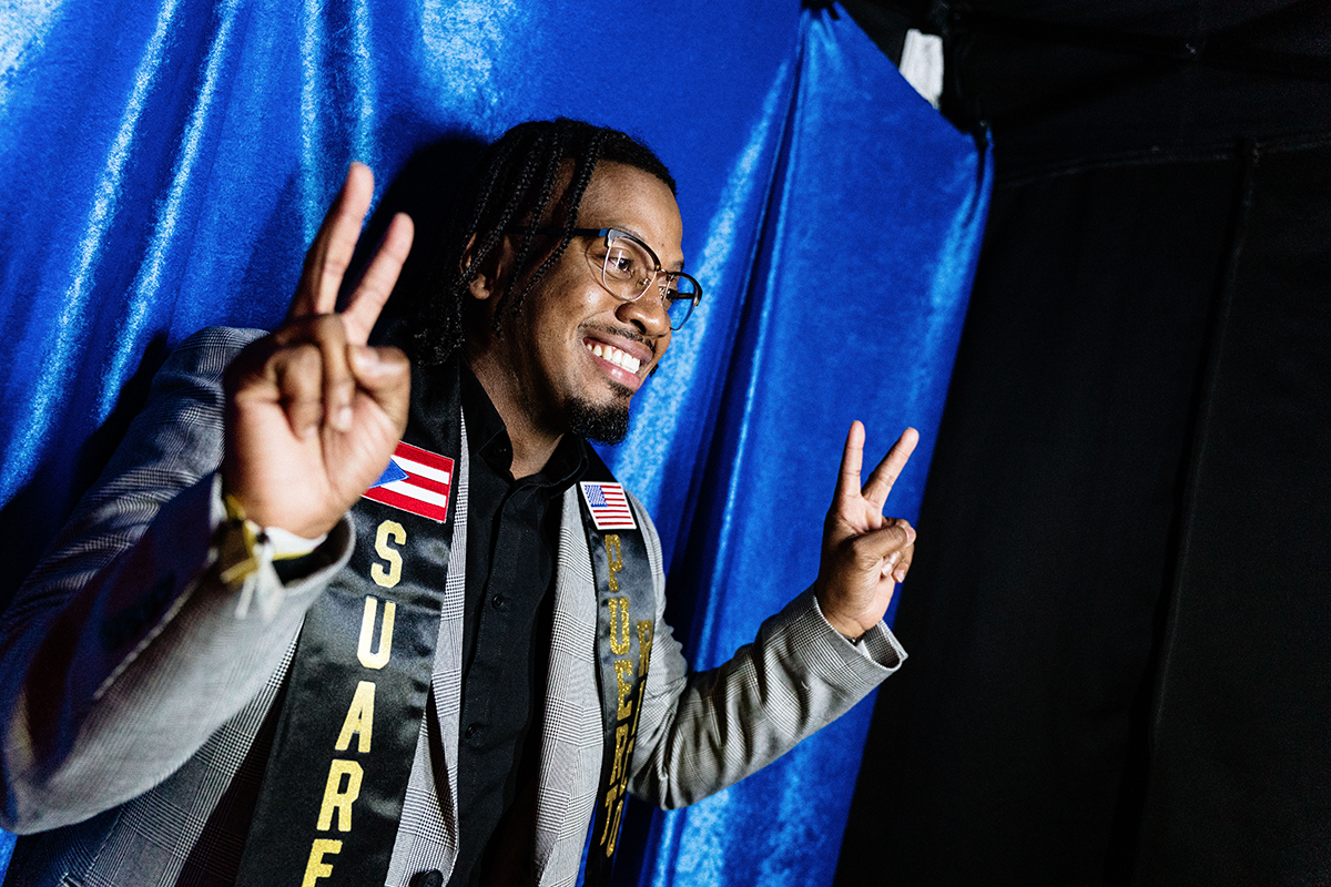 FLINT_Latin Graduation Celebration 2026_0048 Person wearing a gray checkered blazer and a black sash with Puerto Rican and American flags, making peace signs with both hands against a blue backdrop.