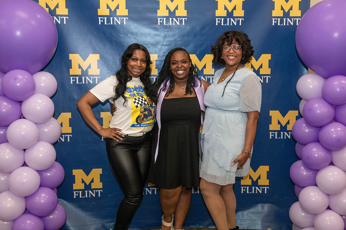 FLINT_Lavender Graduation Celebration 2026_0018 Three women standing in front of a blue backdrop with yellow "M Flint" logos, flanked by purple and lavender balloon columns.