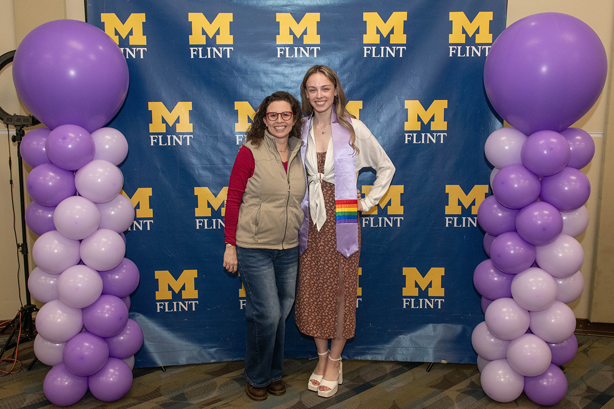 FLINT_Lavender Graduation Celebration 2026_0019 Two people standing in front of a blue backdrop with yellow "M Flint" logos, flanked by purple and lavender balloon columns.