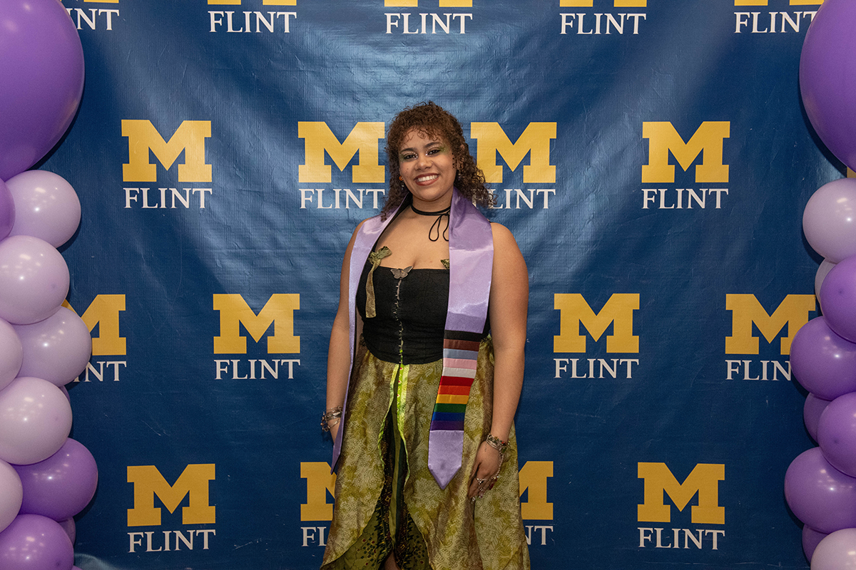 FLINT_Lavender Graduation Celebration 2026_0020 Person wearing a graduation stole with rainbow stripes standing in front of a blue backdrop with yellow "M FLINT" logos and purple balloons on each side.