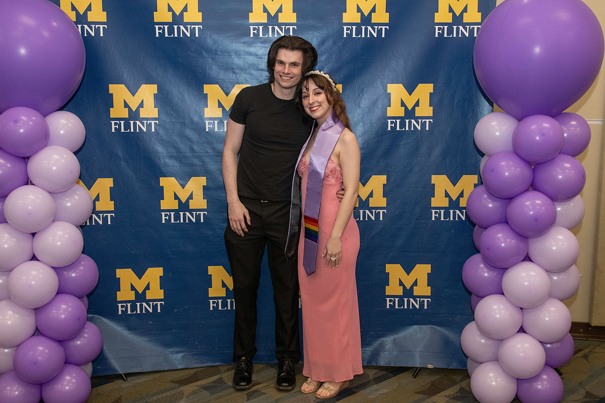 FLINT_Lavender Graduation Celebration 2026_0021 Two people standing in front of a blue backdrop with yellow "M Flint" logos, flanked by purple and lavender balloon columns.