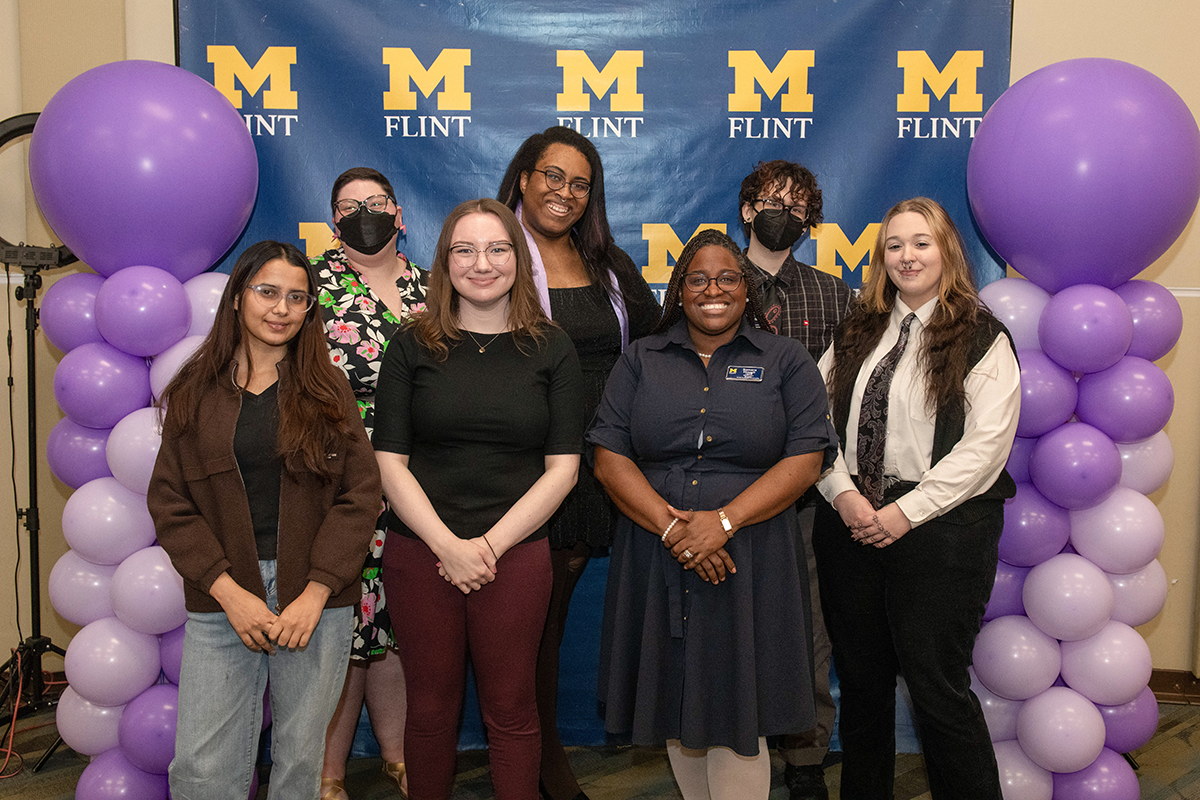FLINT_Lavender Graduation Celebration 2026_0023 Group of seven people posing in front of a blue backdrop with yellow "M Flint" logos and purple balloon columns on each side.