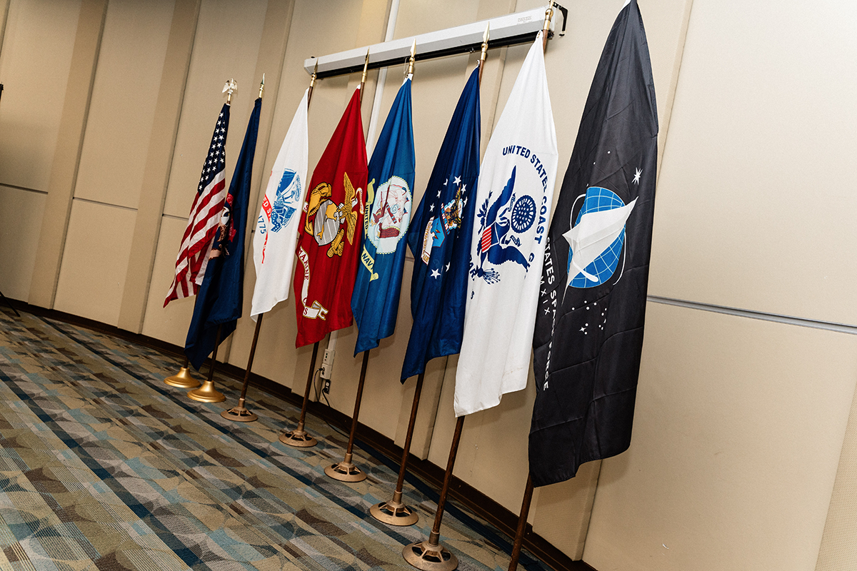 FLINT_Veterans Graduation Celebration 2026_0001 Row of seven U.S. military and government flags standing indoors against a beige wall.