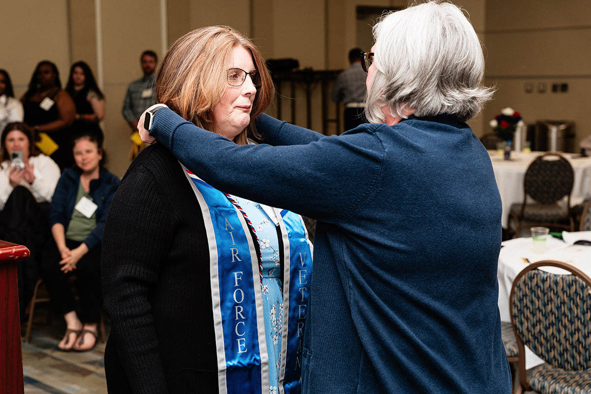FLINT_Veterans Graduation Celebration 2026_0017 A person places an Air Force Veteran sash on another person during an indoor event with seated attendees in the background.