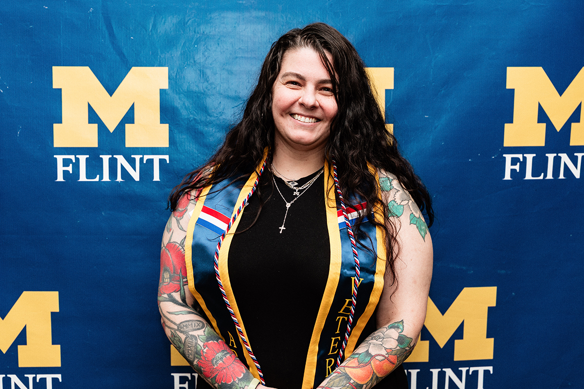 FLINT_Veterans Graduation Celebration 2026_0030 Person with long dark hair and tattoos on both arms wearing a black top and a blue and gold graduation stole standing in front of a blue backdrop with yellow "M FLINT" logos.