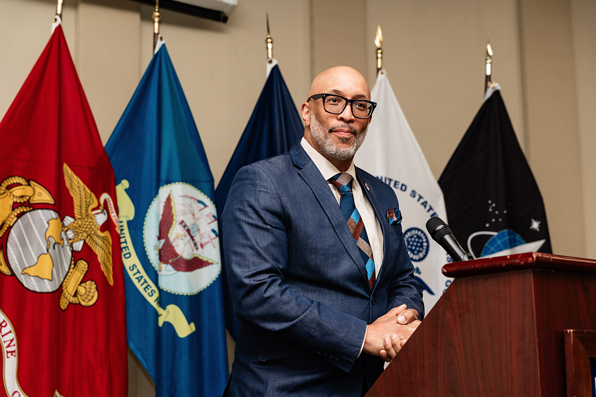 FLINT_Veterans Graduation Celebration 2026_0035 A man in a blue suit and patterned tie standing at a podium with a microphone, with five military flags in the background.