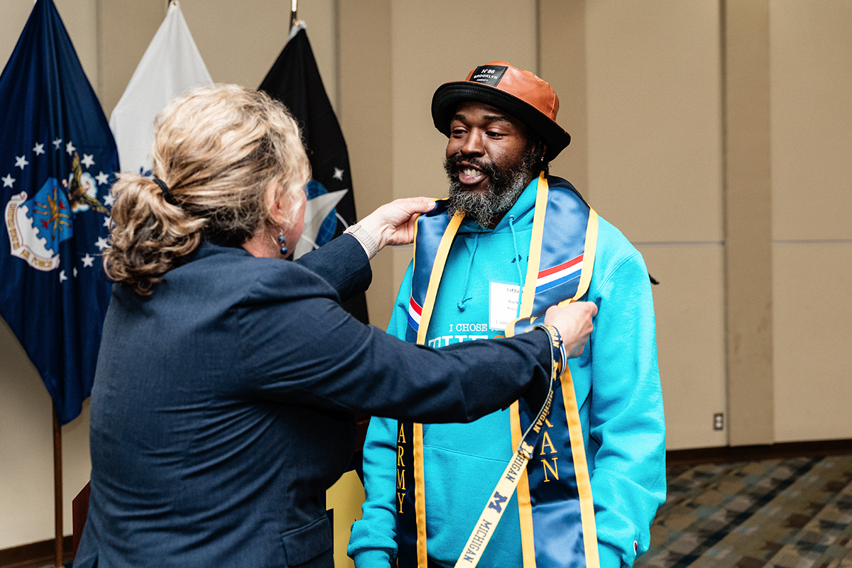 FLINT_Veterans Graduation Celebration 2026_0043 A woman in a dark blazer places a blue and gold Army veteran sash on a man wearing a turquoise hoodie and an orange and black hat, with flags in the background.