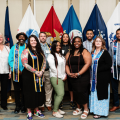 Group of people standing in front of U.S. military service flags including Army, Marine Corps, Navy, Air Force, Coast Guard, and Space Force flags.
