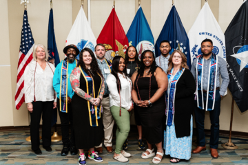 Group of people standing in front of U.S. military service flags including Army, Marine Corps, Navy, Air Force, Coast Guard, and Space Force flags.