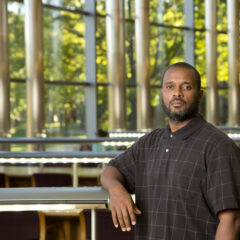 Man in a dark checkered polo shirt leaning on a railing inside a building with large windows and trees visible outside.