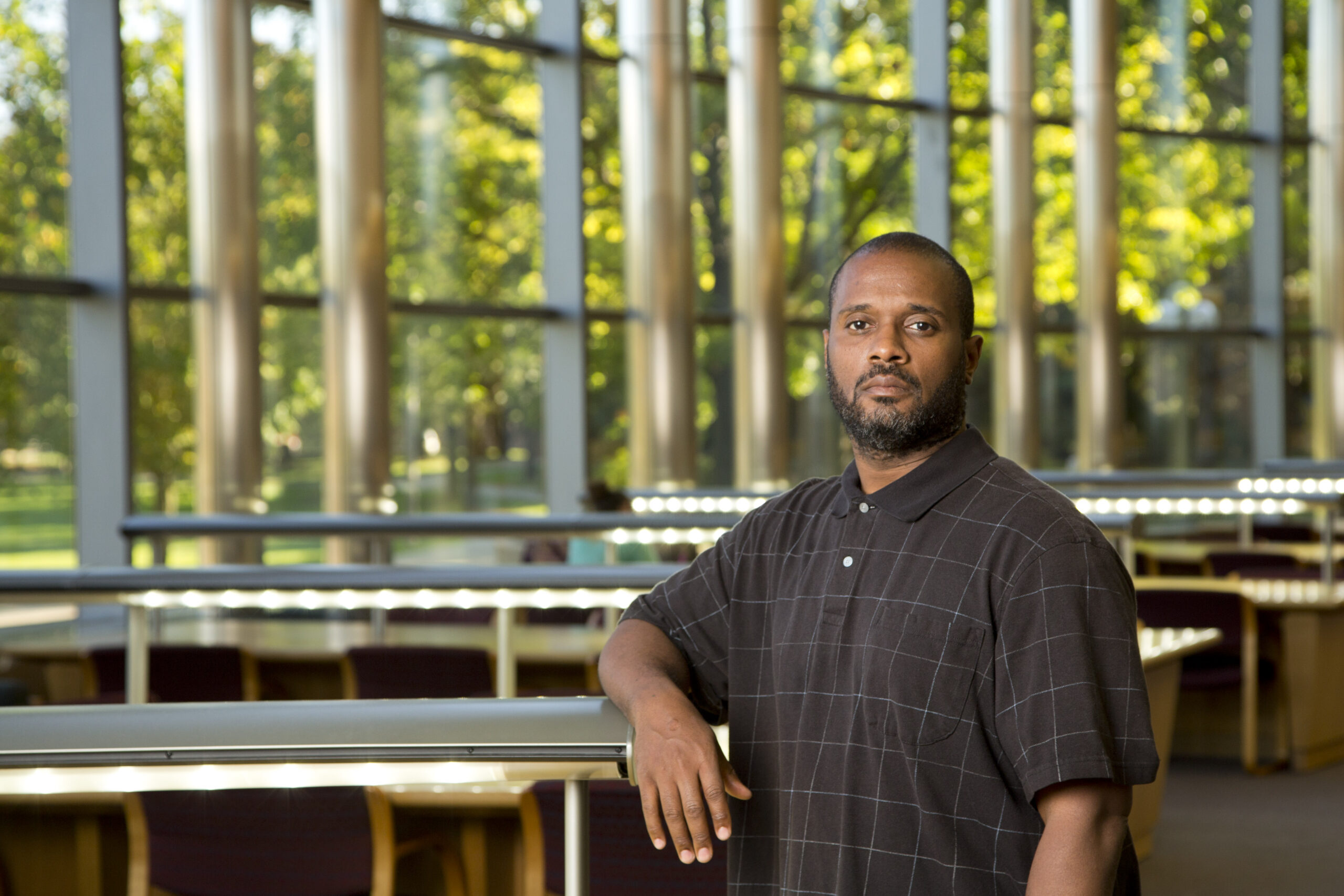 Man in a dark checkered polo shirt leaning on a railing inside a building with large windows and trees visible outside.
