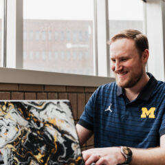 A person wearing a blue University of Michigan polo shirt working on a laptop with a black, white, and gold marbled case, sitting by a window.