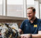 A person wearing a blue University of Michigan polo shirt working on a laptop with a black, white, and gold marbled case, sitting by a window.